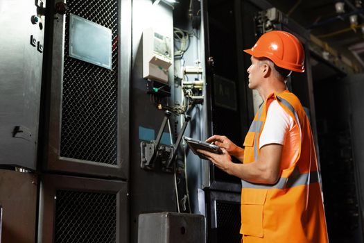 An electrical station engineer in hard hat and vest walks over to a rack and using digital tablet inspect electric power system in electrical control room. Concept of industry, factory, energy.