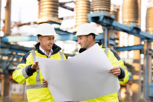 Two engineers are talking in front of electrical transmission lines, working on renewable energy development. Engineers in special clothing discuss a drawing on paper near high-voltage power line.
