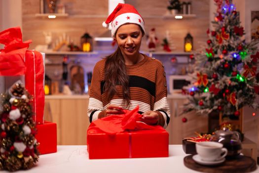 Festive joyful woman with santa hat using ribbon for bow on christmas present box in festive kitchen. Young caucasian adult preparing gift with red wrapping paper for celebration party