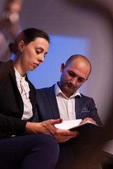 Businessman doing overtime with businesswoman late night afterhours tired on a deadline. Colleague entrepreneur working late together in evening at corporate job sitting on staircase, explaining difficult finance project.