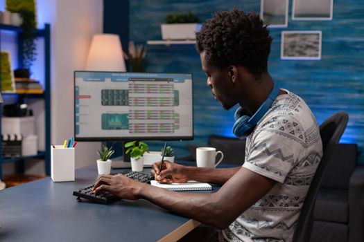 African american young trader writing crypto earning chart on notebook while analyzing stock market investment on computer. Broker man checking marketing trading charts working remote from home