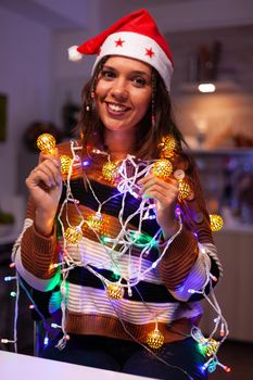 Cheerful adult tangled in christmas string lights used for decorating home. Festive caucasian young woman using garland with illuminated bulbs for seasonal decor and winter tree ornaments