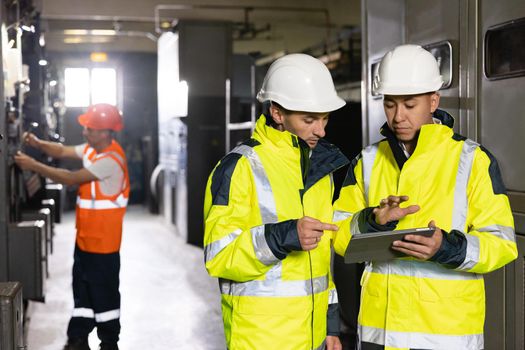 Male Electrical Engineers in Hard Hats Discuss New Project while Using Tablet Computer. They're Making Calculated Engineering Decisions. They Work at the Electric Power Factory.