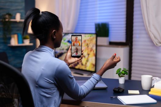 Dark skinned woman using smartphone late night in home office discussing wtih people in video conference. Busy employee using modern technology network wireless doing overtime for job.