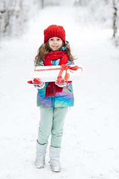 Child in red hat with Christmas presents and gifts in snow. Winter outdoor fun. Kid play in snowy park on Xmas eve. Happy Little caucasian girl smile and holding gift box