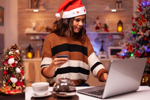 Joyful adult doing online shopping for christmas family dinner at home with seasonal decorations, lights and ornaments. Caucasian woman doing credit card payment for celebration party