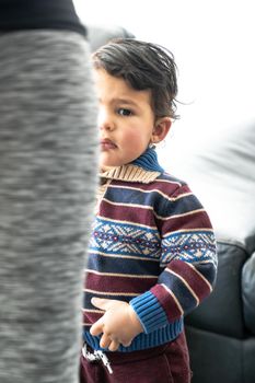 A handsome young baby boy or toddler with long curly brown hair wearing a patterned and striped collared sweater stands by his mother's leg as she combs and fixes the child's hair.