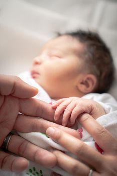 A newborn infant baby girl in a blanket swaddle wraps her tiny hand and fingers around her father and mother's fingers as she sleeps peacefully.