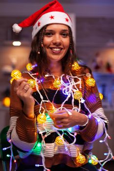 Cheerful adult tangled in christmas string lights used for decorating home. Festive caucasian young woman using garland with illuminated bulbs for seasonal decor and winter tree ornaments