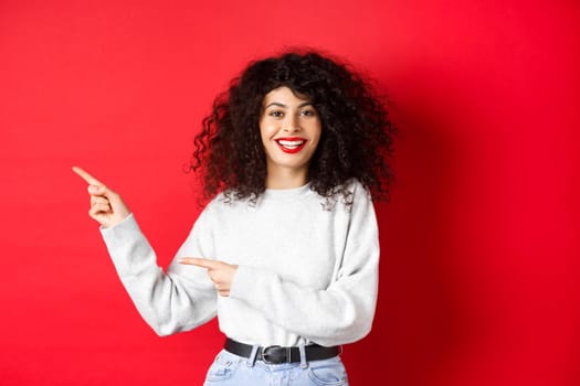 Smiling caucasian woman with curly hair and red lips, pointing fingers left at logo, showing promo offer, standing against studio background.