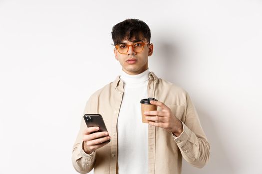 Real people. Stylish hipster guy in glasses holding cup of coffee from cafe takeaway and mobile phone, standing on white background.