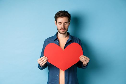 Lonely guy looking sad at valentines red heart with sad face, standing over blue background.