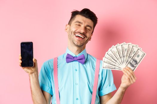 E-commerce and shopping concept. Rich and happy guy laughing, showing money in cash and empty smartphone screen, standing on pink background.