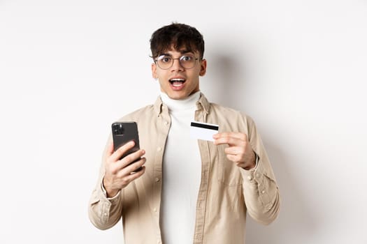 Excited young man shopping online, holding mobile phone and plastic credit card, making purchase in internet, standing on white background.