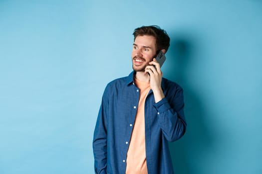 Handsome caucasian man talking on mobile phone, looking aside at empty space with carefree smile, standing on blue background.