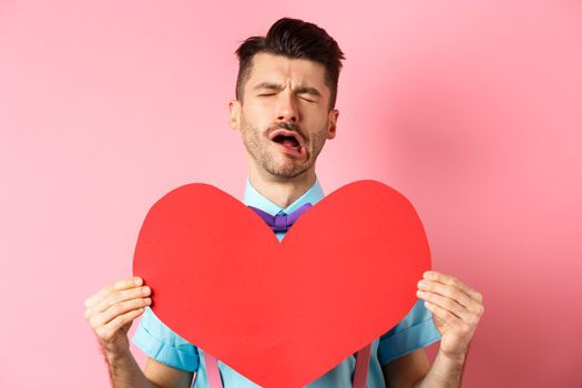 Valentines day concept. Sad and lonely man feeling heartbroken, being rejected, showing big red heart cutout and crying from break-up, standing on pink background.