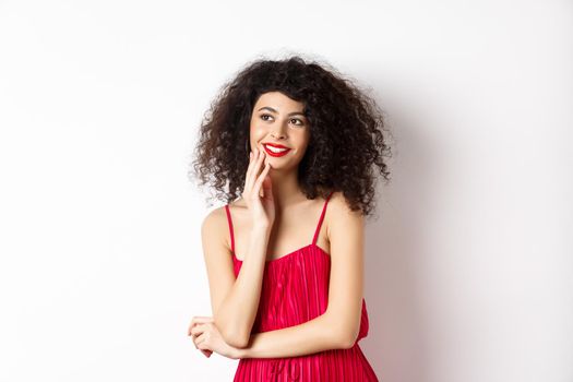 Dreamy elegant girl with curly hair, wearing red dress and makeup, looking left and smiling at logo, standing on white background.