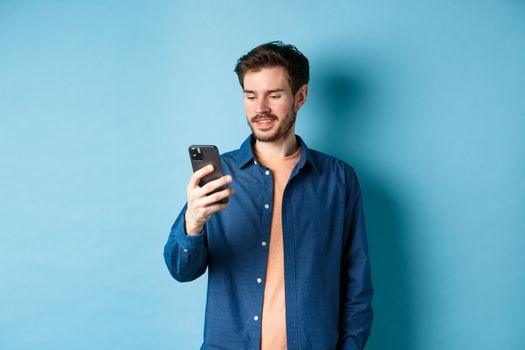 Handsome modern guy using mobile phone, reading smartphone screen and smiling, networking on blue background.