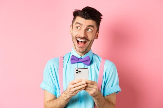 Technology concept. Excited man looking aside with dreamy face, winning online prize on smartphone, standing cheerful over pink background.