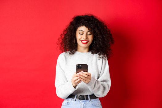 Smiling european woman reading mobile phone screen, texting a message on smartphone, standing in sweatshirt against red background.