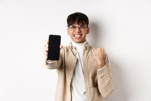Lucky young man winning prize online, showing smartphone screen and rejoicing of good news, standing happy on white background.