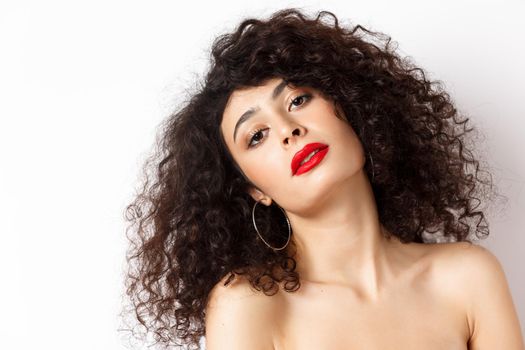 Close-up portrait of sensual and romantic woman with curly hair, red lips, looking seductive at camera, posing on white background.