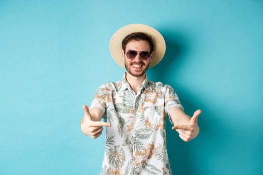 Happy tourist in summer hat and hawaiian shirt, pointing fingers at logo on center, showing something, standing on blue background.