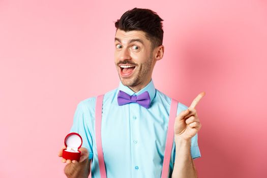 Valentines day. Funny guy with moustache and bow-tie, showing engagement ring and pointing finger at upper right corner, standing on pink background.