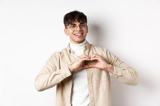 Happy Valentines. Handsome young man in glasses say I love you, showing heart sign and smiling at girlfriend, celebrating lovers day, standing over white background.