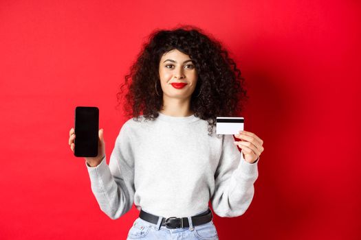 Young modern woman with curly hair showing plastic credit card and mobile phone screen, demonstrating online shopping app, standing on red background.