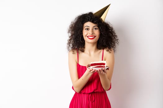 Beautiful woman in red dress, wearing party hat and celebrating birthday, holding b-day cake and making wish, smiling at camera, standing on white background.