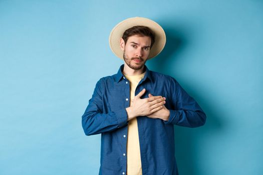 Heartfelt young woman in tourist hat, holding hands on heart and looking thankful and serious, standing on blue background.