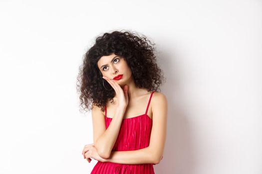 Annoyed and bored young woman with curly hair, look away distressed, lean face on hand, standing bothered in red dress on white background.