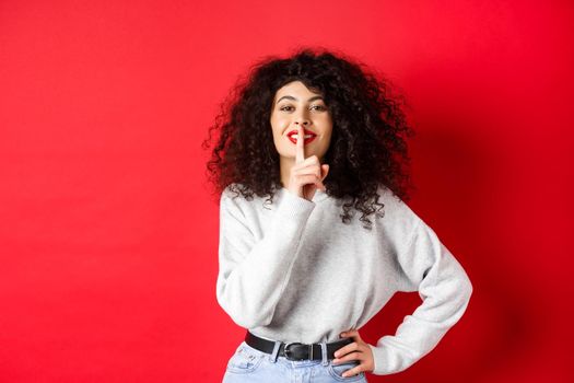 Beautiful modern woman with curly hair and makeup, shushing and smiling, telling a secret, making surprise and smiling, standing over red background.