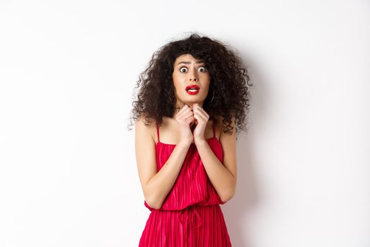 Scared caucasian woman trembling from fear, wearing red dress and staring anxious at camera, standing over white background.