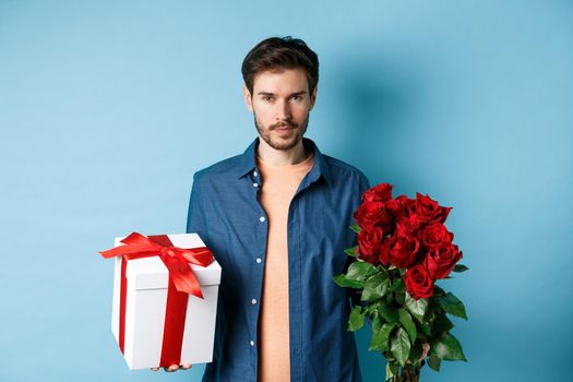 Love and Valentine day concept. Passionate man looking confident at camera, holding gift box and red roses for romantic date, standing over blue background.