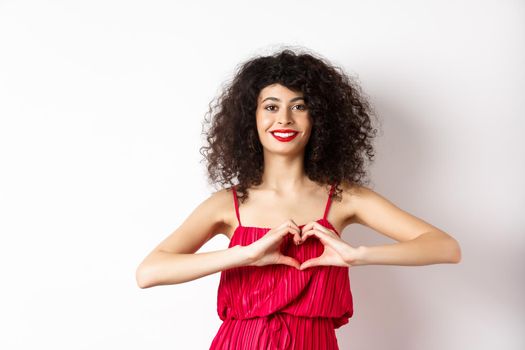 Charming young woman with red romantic fress, showing heart gesture and smiling, express love, standing over white background.