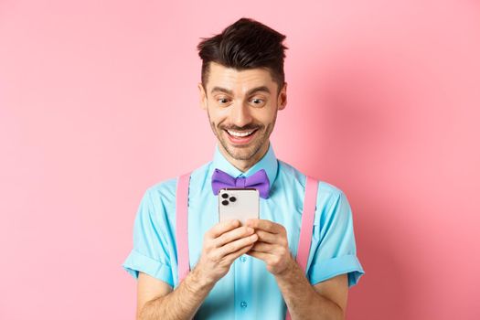 Online shopping. Happy guy looking at smartphone screen, reading message and smiling, standing on pink background.