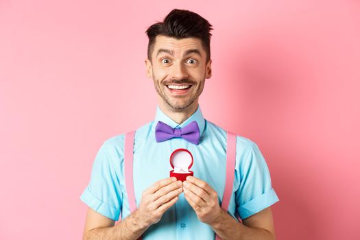 Valentines day. Romantic man in bow-tie showing engagement ring and smiling, asking to marry him, making proposal to lover on pink background.