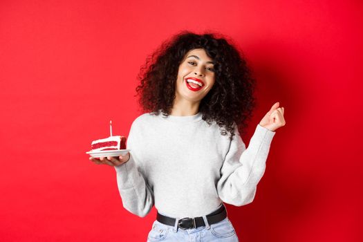 Celebration and holidays concept. Happy beautiful woman dancing and making birthday wish, holding b-day cake and smiling, standing on red background.