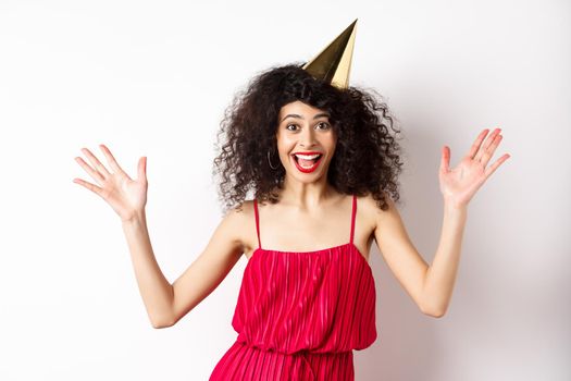 Cheerful young woman in red dress, celebrating birthday, wearing party hat and smiling, screaming of joy, standing on white background.