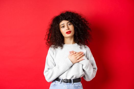 Beautiful woman with curly hair feeling touched and thankful, holding hands on heart and looking with affection at camera, red background.