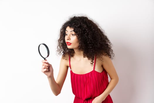 Beautiful woman in red dress searching for something, looking aside through magnifying glass, standing against white background.