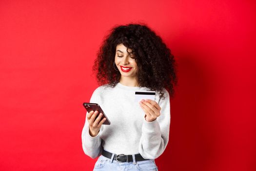 E-commerce and online shopping concept. Attractive caucasian woman paying for purchase in internet, holding smartphone and credit card, red background.