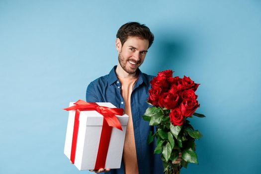 Love and Valentines day concept. Romantic smiling man giving you gift box and bouquet of flowers on date, standing over blue background.