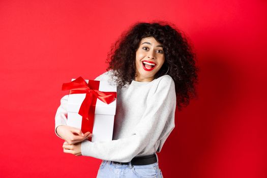 Valentines day. Happy girlfriend holding gift box from secret admirer, looking romantic at lover, receive present, standing on red background.