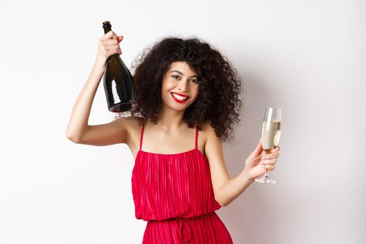 Happy woman partying on valentines day holiday, dancing with glass and bottle of champagne, wearing red dress, smiling on white background.