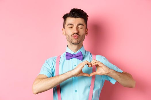 Romantic man in funny bow-tie waiting for kiss, close eyes and pucker lips kissing, showing heart gesture, being in love on Valentines day, standing over pink background.