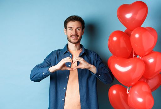 Happy valentines day. Boyfriend in love showing heart gesture to lover and smiling, standing near red balloons on blue background.