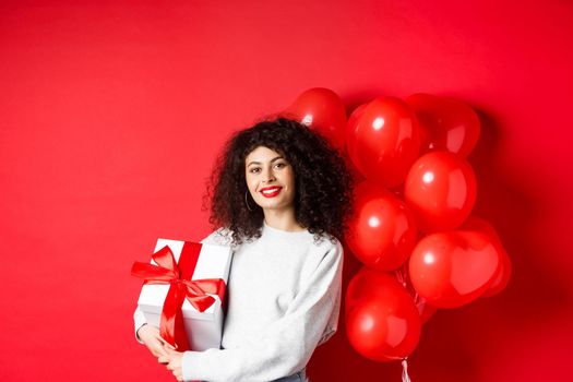 Beautiful caucasian woman with dark curly hair and red lips, standing with romantic hearts balloons and present in box, receive gift from lover, standing on studio background.